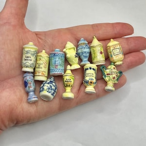 May include: A collection of twelve miniature ceramic apothecary jars and vases in various shapes and sizes. Each piece features hand-painted floral motifs, decorative patterns, and black text labels in shades of blue, yellow, and white.