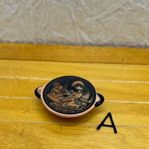 May include: A small, round, black and peach-colored decorative bowl with a detailed illustration of two figures. The figures are in a classical style, with one figure reclining and the other standing. The bowl has two small handles.