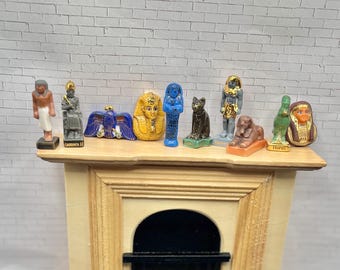 May include: A collection of miniature Egyptian figurines, including pharaohs, cats, and sphinxes, displayed on a wooden shelf. The figurines are painted in various colors, such as gold, blue, and brown, and are arranged in a row. The shelf is part of a decorative fireplace.
