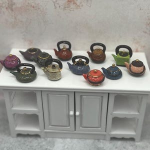 May include: A collection of miniature ceramic teapots in various colors, including green, red, blue, and gold, with black handles. The teapots are arranged on a white cabinet, showcasing a variety of designs and styles.