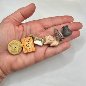 May include: A collection of six small, prehistoric-themed miniature objects including a circular tan disc with an animal carving, an orange rectangular plaque, a tan horse figurine, a tan lion figurine, a dark brown arrowhead, and a small grey pot.