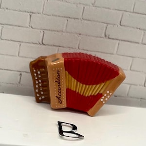 May include: A miniature accordion with a red bellows and tan accents. The word "Accord&eacute;on" is printed on the side. The instrument has white dot details and a gold-colored trim. The accordion is set against a white brick background.