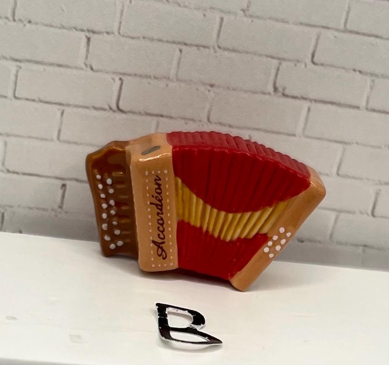 May include: A miniature accordion with a red bellows and tan accents. The word "Accord&eacute;on" is printed on the side. The instrument has white dot details and a gold-colored trim. The accordion is set against a white brick background.