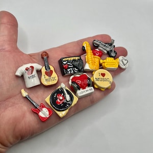 May include: A collection of miniature music-themed ceramic figurines. Includes a white t-shirt with a red heart, guitars, a record player, a saxophone, a trumpet, and a motorcycle. The figurines are in various colors, including red, yellow, black, and white.