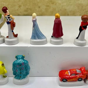May include: A collection of small, colorful figurines featuring various cartoon characters. The figurines include Buzz Lightyear, Woody, Elsa, and others. The figurines are on white bases and appear to be made of ceramic or a similar material.