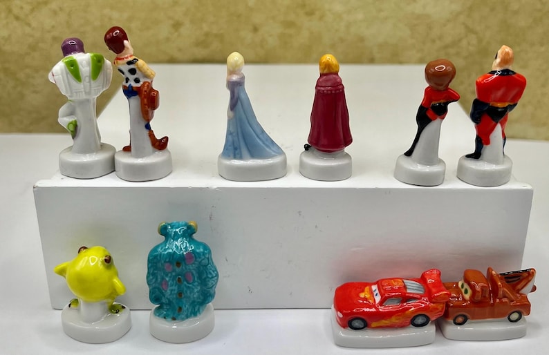 May include: A collection of small, colorful figurines featuring various cartoon characters. The figurines include Buzz Lightyear, Woody, Elsa, and others. The figurines are on white bases and appear to be made of ceramic or a similar material.