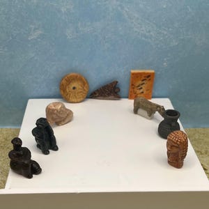 May include: A collection of small, carved figurines and objects displayed on a white surface. The items include human figures, animals, and abstract shapes in various colors, including brown, black, and beige. The background is a light blue wall.