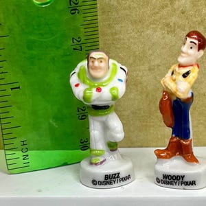 May include: Two small ceramic figurines of Disney Pixar characters Buzz Lightyear and Woody. Buzz is in a white and green spacesuit, and Woody is in a cowboy outfit. A green ruler is in the background, measuring in inches.