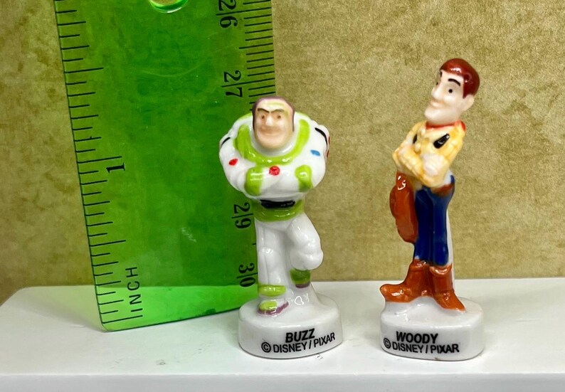 May include: Two small ceramic figurines of Disney Pixar characters Buzz Lightyear and Woody. Buzz is in a white and green spacesuit, and Woody is in a cowboy outfit. A green ruler is in the background, measuring in inches.