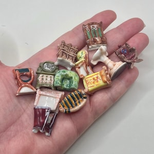 May include: A collection of miniature ceramic furniture pieces, including chairs, a clock, and a table, in various colors and designs. The pieces are held in a hand, showcasing their small size and intricate details. The collection includes a variety of styles.