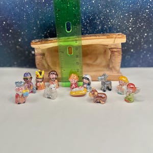 May include: A miniature ceramic nativity scene with figures including Mary, Joseph, baby Jesus, angels, and the Three Wise Men. The figures are colorful and detailed, with a wooden stable in the background. A green ruler is placed next to the figures.