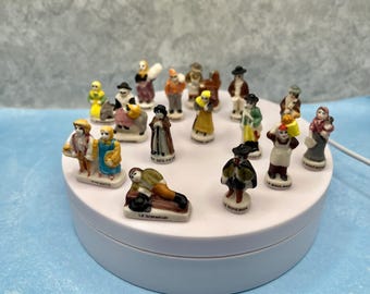May include: A collection of small, hand-painted porcelain figurines depicting various figures in traditional attire. The figurines are arranged on a white rotating platform against a light blue backdrop. Each figurine is approximately 2 inches tall.