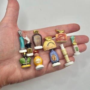 May include: A collection of eight miniature ceramic perfume bottles in various shapes and colors, including blue, yellow, green, and gold accents. Each small decorative vessel features unique detailing and a glossy finish, displayed together.