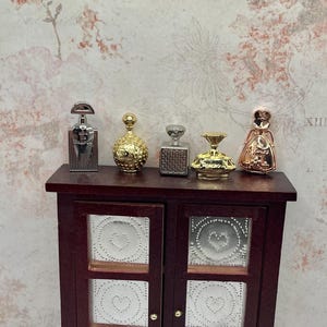 May include: Miniature perfume bottles in silver, gold, and copper tones are displayed on a dark wood cabinet. The bottles have various shapes and designs, including square, round, and ornate styles. The cabinet has two doors with decorative metal inserts.