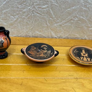 May include: A collection of miniature Greek-style pottery. Includes a small vase with handles, a shallow dish with handles, and a round plate. The pieces feature black and orange painted figures on a cream background.
