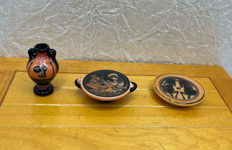 May include: A collection of miniature Greek-style pottery. Includes a small vase with handles, a shallow dish with handles, and a round plate. The pieces feature black and orange painted figures on a cream background.