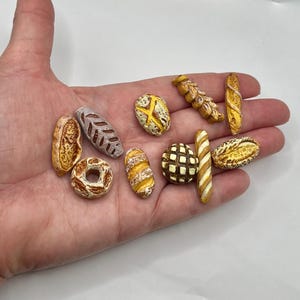 May include: A collection of miniature bread items, including a donut, baguette, and various loaves, in shades of brown and yellow. The assortment is held in a hand, showcasing the detailed textures and shapes of each baked good. The miniature food items are likely for a dollhouse or craft project.