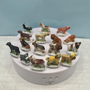 May include: A collection of small, ceramic dog figurines in various breeds and colors, displayed on a white rotating platform. Each figurine is detailed with realistic features and rests on a small base.