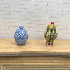 May include: Two miniature ceramic vases. One is light blue with a detailed embossed pattern. The other is a colorful, patterned vase with yellow, green, red, and pink accents. The vases are displayed on a light wood surface.