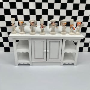 May include: A white wooden cabinet with two doors and open shelves, topped with seven small ceramic chef figurines. The chefs wear white uniforms and hats, holding various kitchen tools. The background features a black and white checkered pattern.