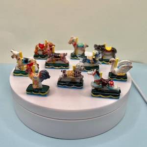 May include: A collection of miniature carousel animal figurines, including a rooster, horses, dogs, and a swan. Each figurine is brightly colored and mounted on a small, decorative base. The carousel animals are arranged on a white rotating platform.