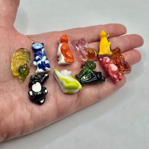 May include: A collection of small, colorful glass figurines in various animal shapes, including cats, bears, and turtles. The figurines are in shades of yellow, orange, blue, green, pink, and black. The figurines are held in a hand.