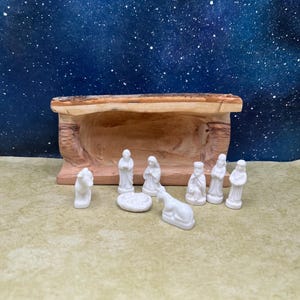 May include: A handcrafted nativity scene featuring a wooden manger and small white porcelain figurines. The set includes figures of Mary, Joseph, the baby Jesus, and other biblical characters, along with animals. The background is a starry night.