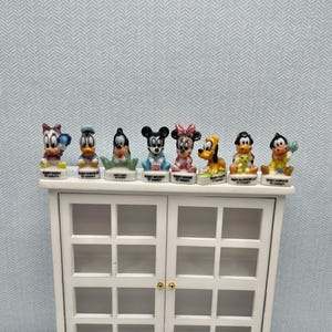 May include: A collection of eight ceramic Disney figurines, including Mickey Mouse, Minnie Mouse, Donald Duck, and Pluto, displayed on a white cabinet. Each figurine is brightly colored and features the character's likeness. The figurines are labeled with the character's name.