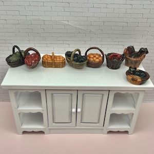 May include: A collection of miniature ceramic baskets filled with various items, including fruit and a cat. The baskets are in shades of brown, orange, and green, arranged on a white table. The table has a cabinet with doors and shelves.