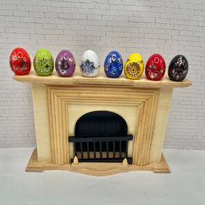 May include: A miniature fireplace mantel with a cream-colored finish, holding seven decorative, hand-painted Easter eggs. The eggs are in various colors, including red, green, purple, white, blue, yellow, and black, each with unique floral designs.