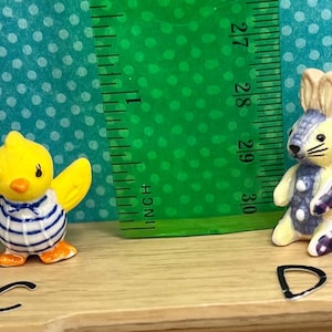 May include: A yellow chick figurine with outstretched wings, wearing a blue and white striped shirt. A bunny figurine with a patchwork design in purple and blue. A green ruler shows measurements in inches.