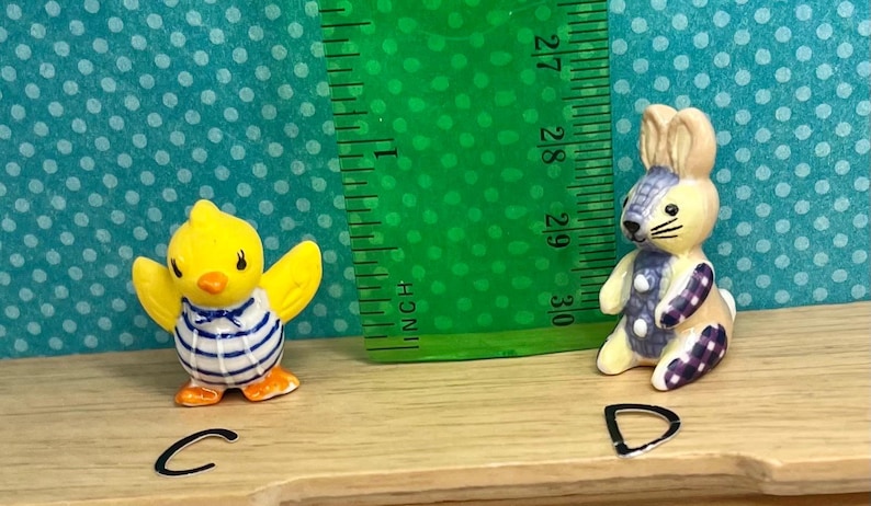 May include: A yellow chick figurine with outstretched wings, wearing a blue and white striped shirt. A bunny figurine with a patchwork design in purple and blue. A green ruler shows measurements in inches.