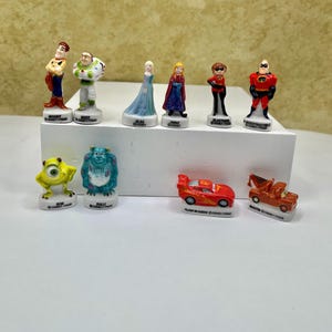 May include: A collection of Disney Pixar figurines, including characters like Woody, Buzz Lightyear, Elsa, and Mr. Incredible. The figurines are brightly colored and stand on small white bases. Also included are figurines of Bob, Sully, Flash McQueen, and Mater.