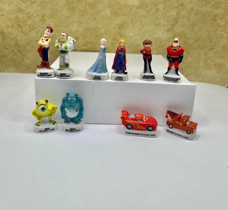 May include: A collection of Disney Pixar figurines, including characters like Woody, Buzz Lightyear, Elsa, and Mr. Incredible. The figurines are brightly colored and stand on small white bases. Also included are figurines of Bob, Sully, Flash McQueen, and Mater.