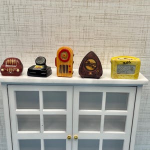 May include: Miniature vintage-style figurines of a car, telephone, radio, clock, and digital clock are displayed on a white cabinet. The figurines are painted in various colors, including red, black, yellow, and brown. The cabinet has glass doors.
