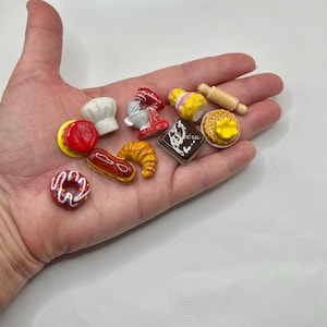 May include: A collection of miniature bakery-themed charms, including a chef's hat, a mixer, a croissant, a donut, and a rolling pin. The charms are brightly colored and detailed, perfect for crafting or jewelry making.
