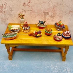 May include: A miniature yellow table displays several small, decorative food items. These include a fish, a dish of vegetables, a cornucopia, and several small cakes. A small white and gold pitcher is also present. The items are colorful and detailed.