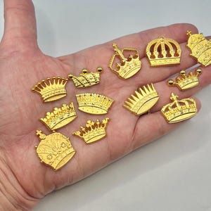 May include: A collection of gold-colored crown charms in various designs. The charms are displayed on a hand, showcasing their intricate details and regal appearance. These decorative charms are suitable for jewelry making or craft projects.