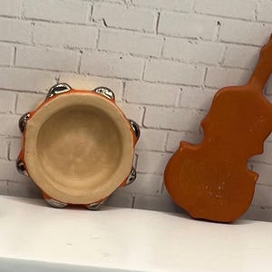 May include: A collection of miniature musical instruments, including a red and tan accordion, a tambourine, a violin, and two drums. The instruments are arranged on a white surface against a white brick wall.