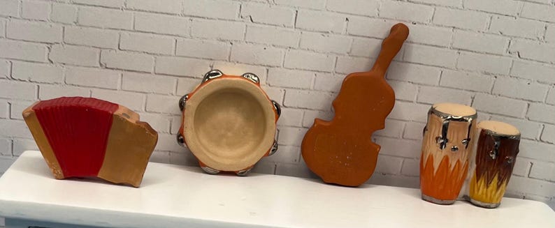 May include: A collection of miniature musical instruments, including a red and tan accordion, a tambourine, a violin, and two drums. The instruments are arranged on a white surface against a white brick wall.
