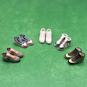 May include: Miniature sneakers in various styles and colors, including white, blue, brown, and green, are arranged on a green surface. The collection features low-top and high-top designs, showcasing a variety of athletic footwear.
