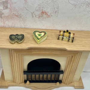 May include: A miniature wooden fireplace mantel featuring a light brown top surface and a black metal grate. Three small decorative trinket boxes are placed on the mantel: one butterfly-shaped, one heart-shaped, and one rectangular chest-style box.