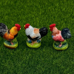 May include: Three ceramic rooster figurines with red combs and colorful plumage. The roosters are perched on green bases and set against a backdrop of green grass. The roosters have detailed feathers and varied color patterns.