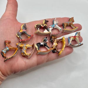May include: A collection of miniature rocking horses in various colors and designs. The horses are painted with details like saddles, manes, and decorative patterns. The rocking horses are small, likely a few inches in size, and are held in a hand.