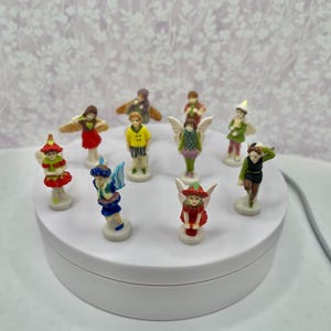 May include: A collection of small fairy figurines in various poses and outfits. The fairies are painted with bright colors, including red, green, yellow, and blue. Some have wings, and all are standing on small white bases.
