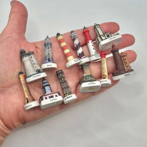 May include: A collection of twelve miniature lighthouse figurines, each featuring distinct architectural designs, color patterns, and labels including Ouessant, Sete, Pas de Calais, Cote d'Armor, Arcachon, Brest, Camargue, Corse, and St Malo, displayed together.