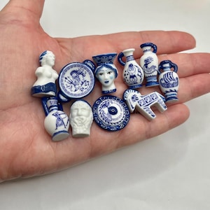 May include: A collection of miniature blue and white ceramic figurines, including vases, plates, and sculptures. The figurines feature classical designs and patterns, held in a hand against a white background. The collection includes a statue, a horse, and a bust.