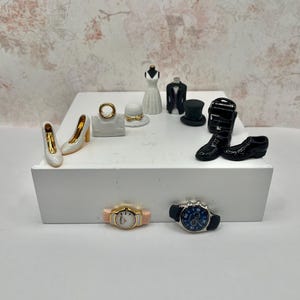 May include: Miniature figurines of fashion accessories, including white high heels with gold accents, a handbag, a ring, a hat, a dress form, a tuxedo, a top hat, a briefcase, black dress shoes, and two watches.