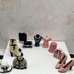 May include: Miniature fashion accessories including a white hat with a black bow, a gray jacket, and beige and black heeled boots. Other items include a black handbag, a necklace, a pink dress, and pink heels.