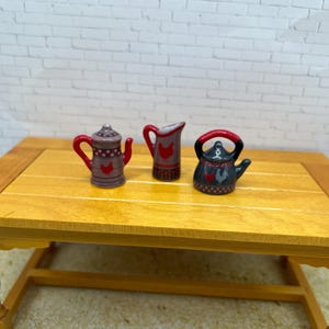 May include: Three miniature ceramic kitchenware pieces: a teapot, a pitcher, and a kettle. The teapot is purple with red accents and a rooster design. The pitcher is red and gray with a rooster. The kettle is black with red accents and a rooster design.